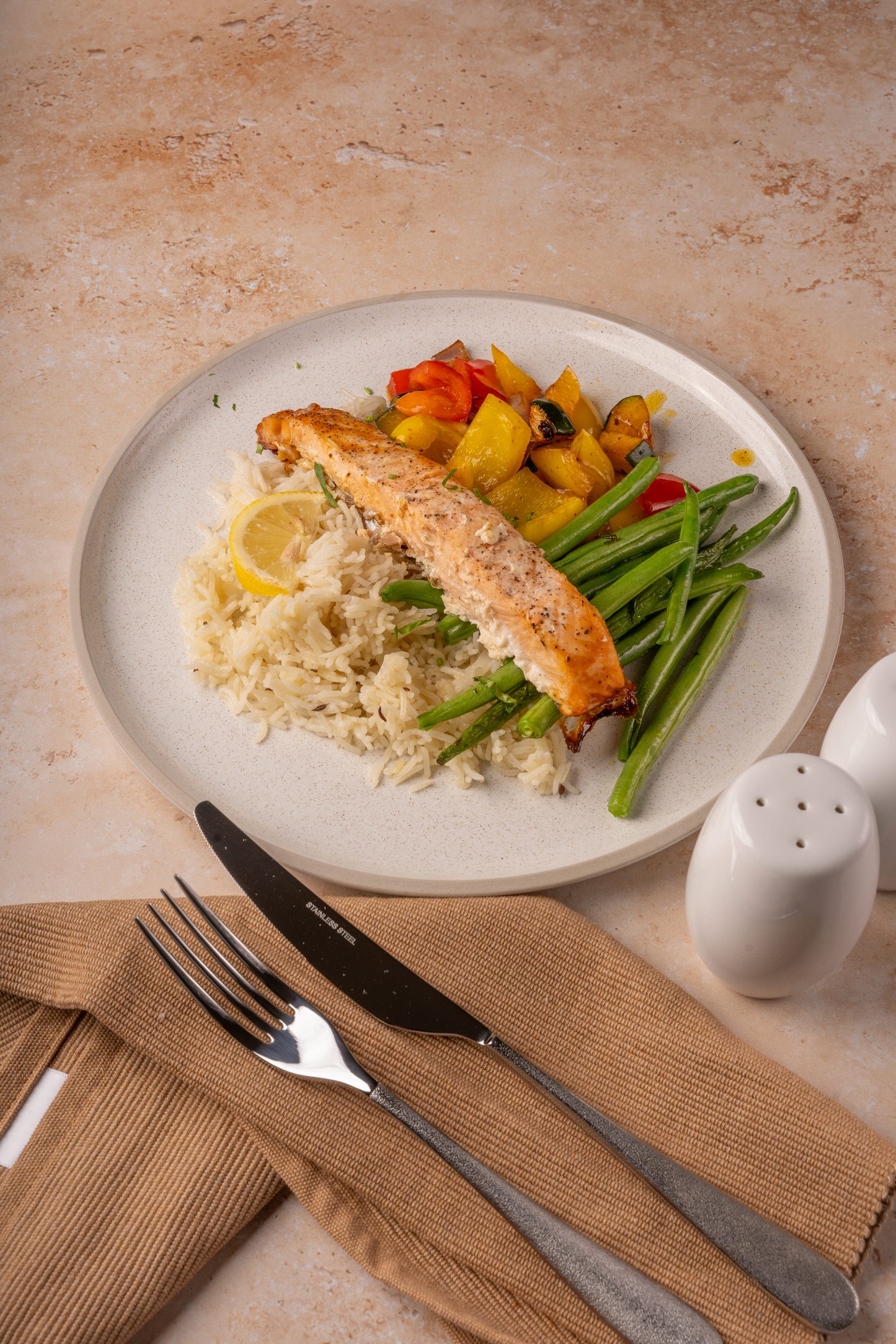 Plated meal with salmon, vegetables, and rice on a beige surface with cutlery and saltshaker.