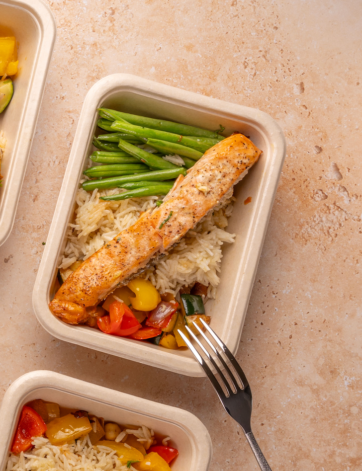 Meal prep containers with rice, vegetables, and slamon on a beige surface.
